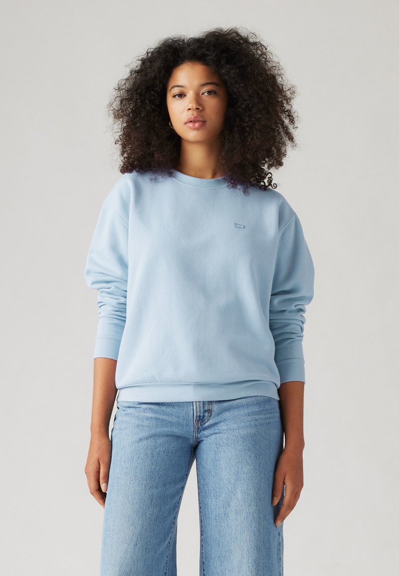 Sweatshirt bleu clair avec un col rond, des poignets et un ourlet côtelés ; tissu doux ; petit logo sur la poitrine, associé à un jean bleu clair.