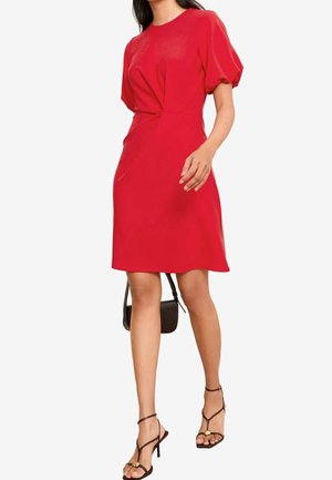 Femme marchant dans une robe rouge mi-longue aux manches bouffantes, portant des talons noirs à brides et un petit sac à main noir.