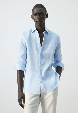 Hombre con camisa de lino azul claro de mangas remangadas y pantalones blancos, de pie frente a un fondo liso y claro.