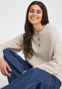 Femme souriante aux longs cheveux foncés portant un pull beige à col et un jean bleu, assise de manière décontractée devant un fond clair et uni.