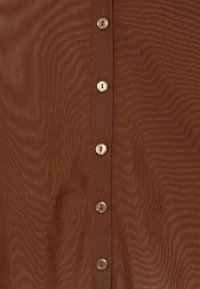 Camisa castanha de botões, com uma textura suave, padrão ondulado e um leve brilho, apresentando seis botões de cor creme na frente.