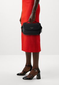 Punaisiin pukeutunut henkilö kantaa pientä mustaa Kate Spade -käsilaukkua ja käyttää mustia kiiltonahkaisia Mary Jane -kenkiä, joissa on nilkkaremmit.