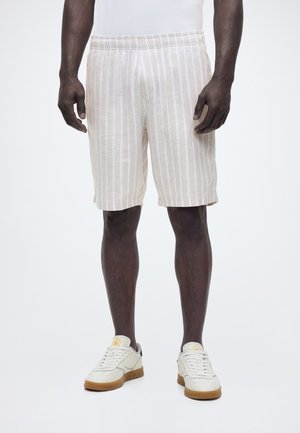 Άνδρας μοντέλο που φοράει βερμούδα μήκους μέχρι το γόνατο με κάθετες ρίγες σε μπεζ και λευκό και λευκά αθλητικά παπούτσια με καουτσούκ σόλες, στέκεται μπροστά σε λευκό φόντο.