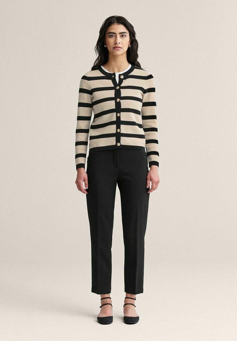 Beige ja mustriline kardigan ümmarguse kaelusega, kullatud nööpidega, koos mustade kohandatud pükste ja mustade nööpidega lamedate kingadega, millel on pahkluu rihmad.