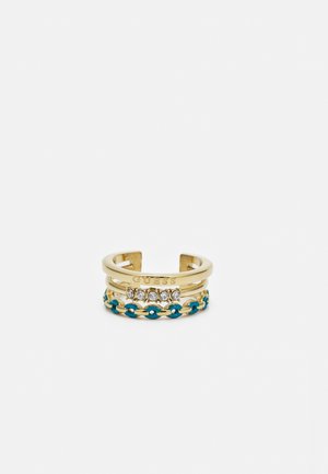 Åben ring i guldfarve med GUESS-logo, fem klare sten og et blåt emalje kædedetaljebånd stablet sammen.
