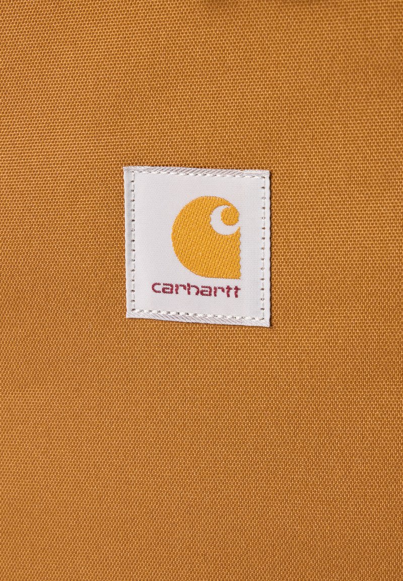 Etiqueta cuadrada de tela blanca con el logo naranja de Carhartt y el nombre de la marca en rojo cosido sobre una tela marrón texturizada.