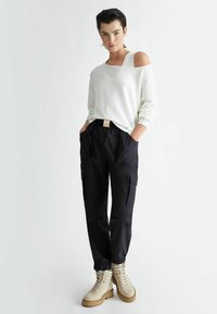 Felpa bianca off-the-shoulder abbinata a pantaloni cargo neri con tasche laterali e polsini elasticizzati, completata da stivali beige con suola spessa.