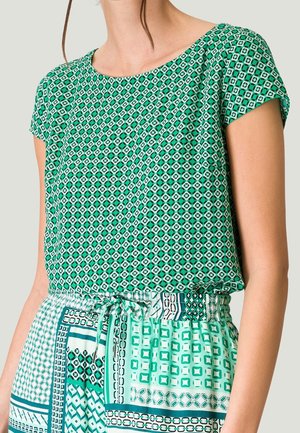 Femme portant un haut à manches courtes vert à motifs et une jupe assortie à motifs avec une taille ajustable par un cordon sur un fond uni.