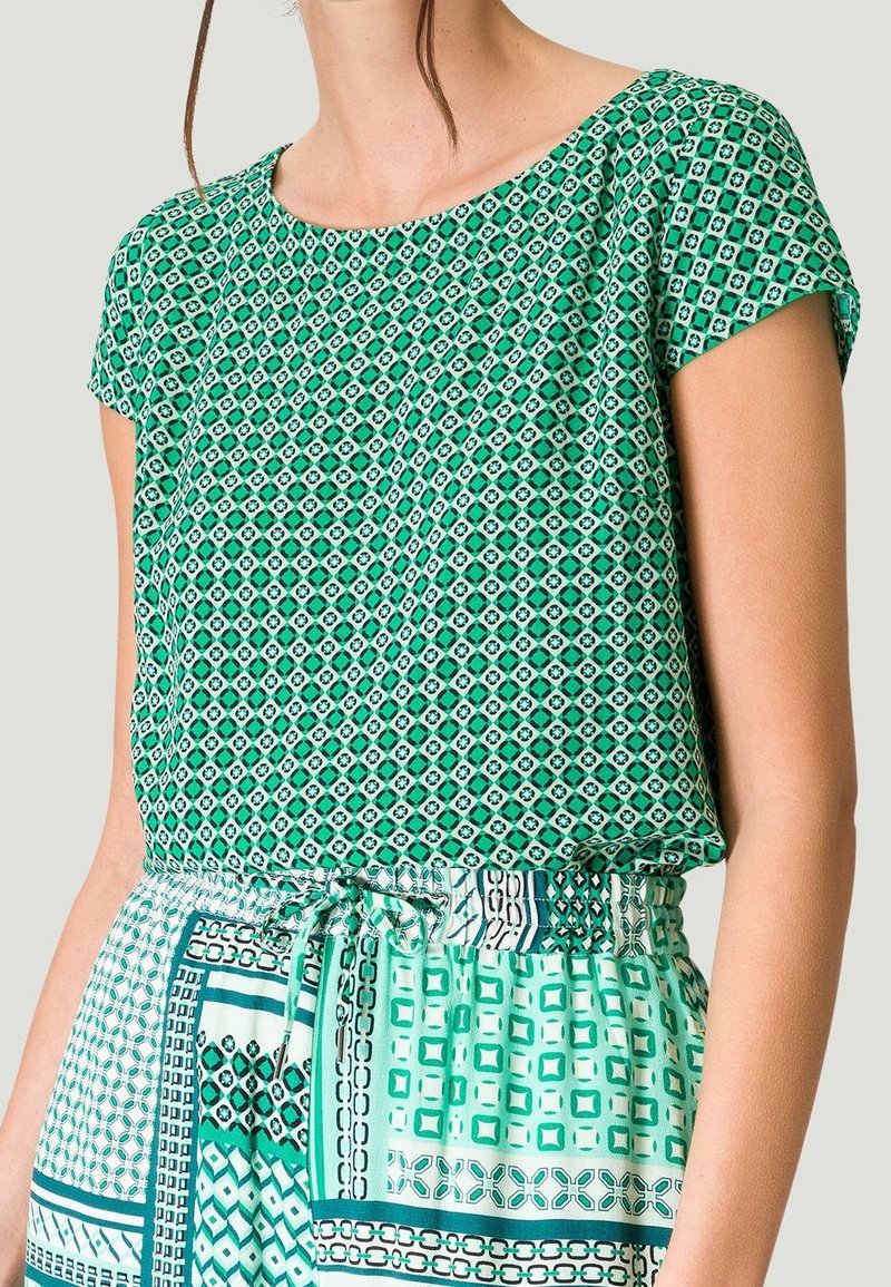 Femme portant un haut à manches courtes vert à motifs et une jupe assortie à motifs avec une taille ajustable par un cordon sur un fond uni.