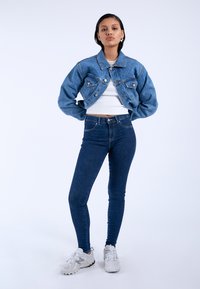 Denim jacket in light blue with button closure and chest pockets, paired with fitted dark blue jeans and white athletic shoes.