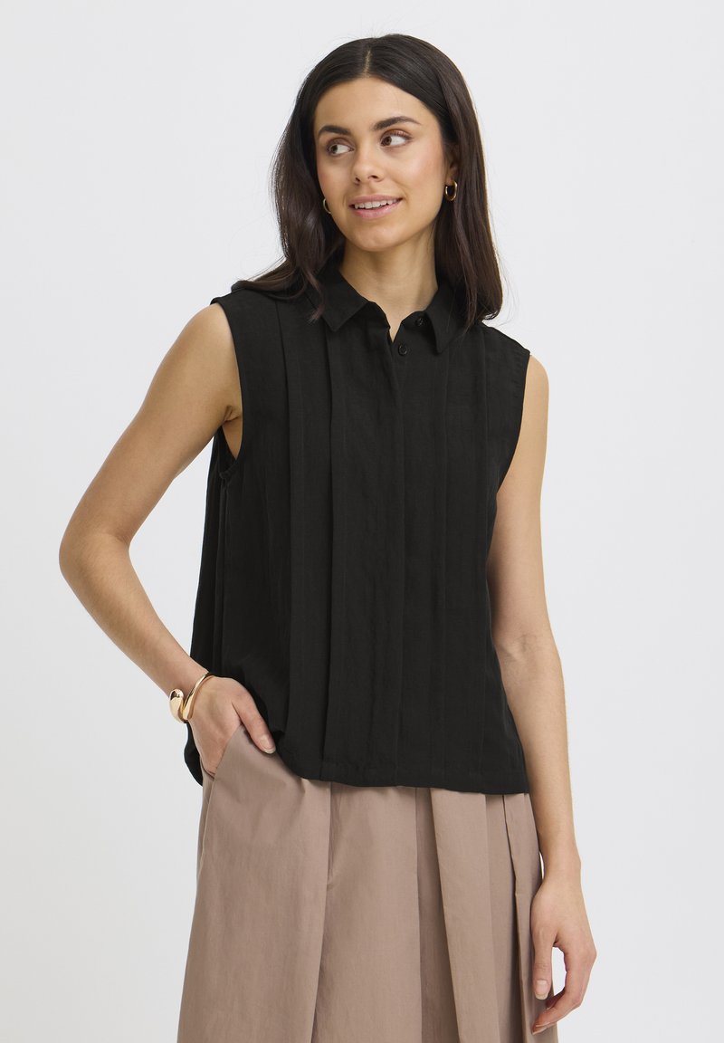 Nainen pukeutuneena mustaan, hihattomaan napitettuun paitapuseroon ja beigen värisiin leveälahkeisiin housuihin, poseeraa käsi taskussa yksiväristä taustaa vasten.