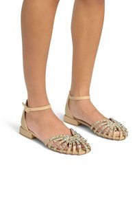 Beige flache Sandalen mit einem Knöchelriemen, die ein Käfigdesign aufweisen, verziert mit hellblauen und klaren Strasssteinen im Zehenbereich.