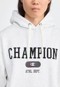 Šedá mikina s kapucňou a čiernym nápisom "CHAMPION", bielymi šnúrkami a kruhovým logom na hrudi. Mäkká textúra materiálu.
