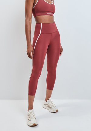 Donna che indossa leggings corti a vita alta color ruggine e reggiseno sportivo abbinato con strisce bianche e scarpe da ginnastica bianche, in piedi contro un muro semplice.