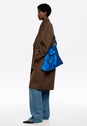 Brązowy oversize'owy płaszcz z dużymi guzikami, niebieska torebka w wzory na ramieniu, szerokie niebieskie dżinsy i czarne buty w szpic.