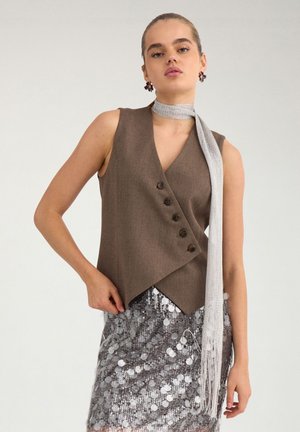 Femme portant un gilet marron sans manches avec des boutons, une jupe argentée à sequins, une écharpe argentée nouée autour du cou et des boucles d'oreilles florales foncées, debout devant un fond uni.