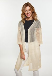 Cardigan lavorato a maglia beige con lavorazione aperta, maniche a tre quarti e vestibilità ampia, indossato sopra una maglietta nera e abbinato a pantaloni bianchi.