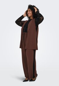Brown matching set featuring a sleeveless vest and loose pants with side black stripes. The top layer has a flowing design, and the model wears black shoes.