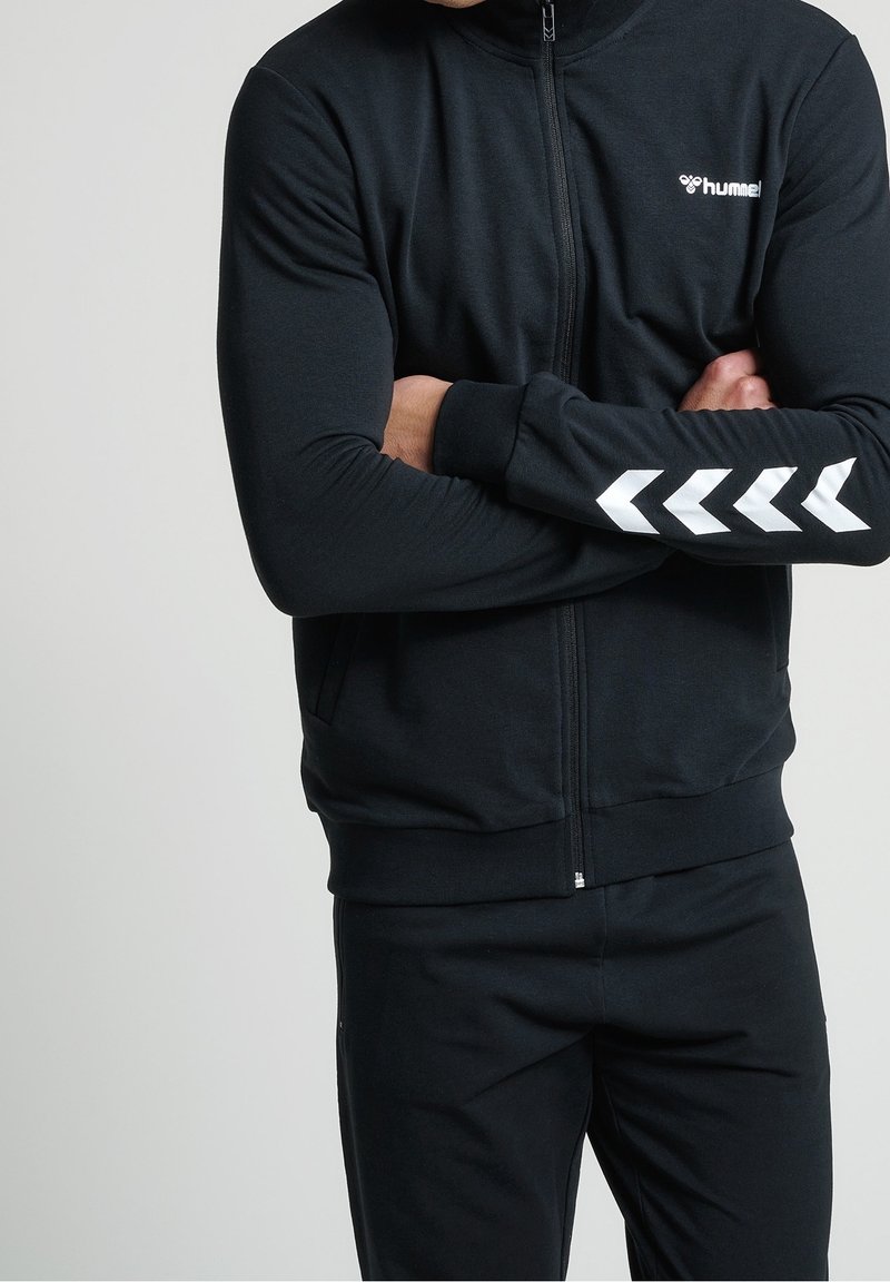 Homme portant une veste de sport noire zippée avec un logo hummel blanc sur la poitrine et des rayures blanches en forme de chevron sur les manches, bras croisés.