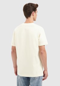 Jonge man in een eenvoudig wit T-shirt met korte mouwen en lichtblauwe jeans, met zijn rug naar de camera gekeerd tegen een lichtgrijze achtergrond.