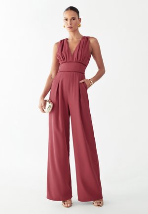 Kobieta ubrana w bezrękawnikowy, głęboki dekolt w kształcie litery V oraz szerokie spodnie w kolorze bordowym, trzymająca małą metaliczną kopertówkę, ozdobiona złotymi kolczykami, bransoletką i sandałami.