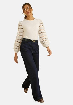 Biely dlhý rukáv s otvoreným čipkovaným vzorom, v kombinácii s tmavými širokými džínsovými nohavicami. Outfit dopĺňajú topánky so zvieracím motívom.