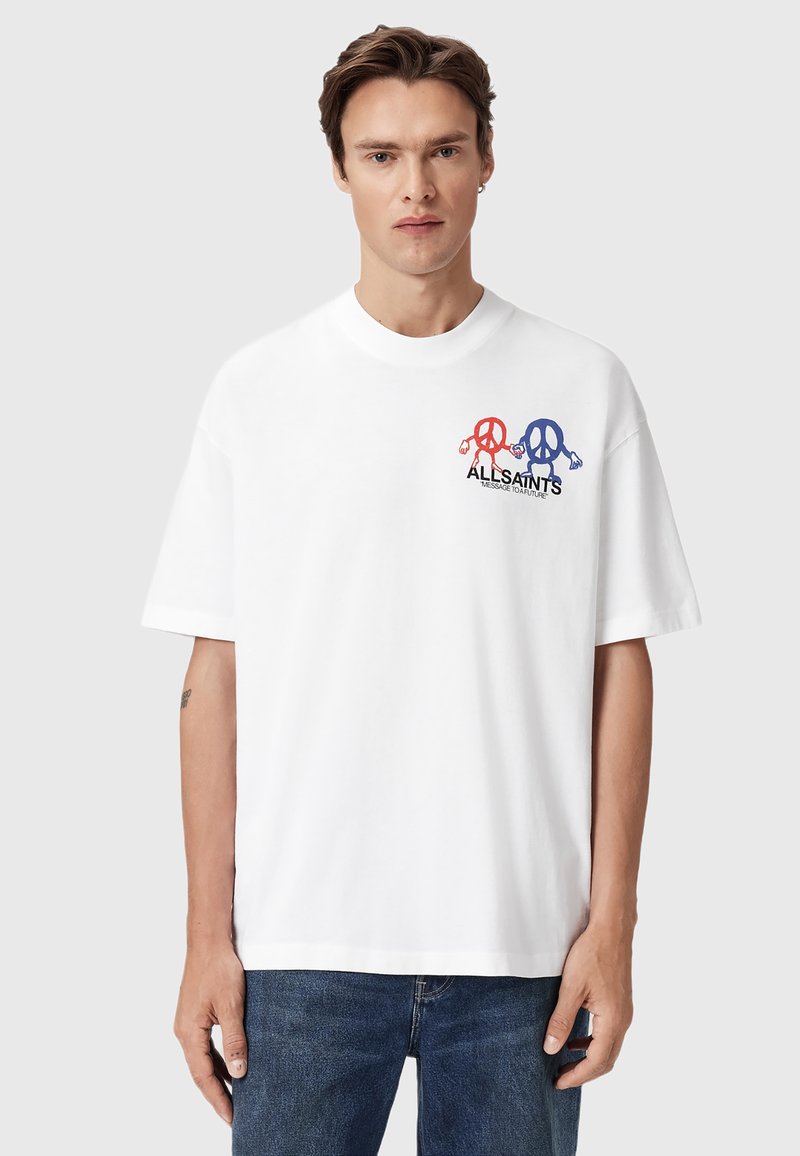 Joven vestido con una camiseta blanca de gran tamaño con personajes de signo de paz en rojo y azul y texto en negro, combinada con jeans azules, de pie contra un fondo liso.