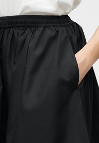 Roka melnas elastīgās vidukļa svārku kabatā, kas kombinēti ar baltu augšu, demonstrējot auduma tekstūru un kabatas detaļas.