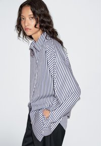 Koszula w paski w kolorach granatowym i białym, o luźnym kroju, z dużym kołnierzem i długimi rękawami zakończonymi podwiniętymi mankietami. Gładka faktura tkaniny.