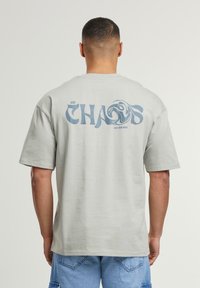 Lys grå bomulds t-shirt med en stor blå tekstgrafik "CHAOS" og et svulmønster på ryggen. Korte ærmer og afslappet pasform.