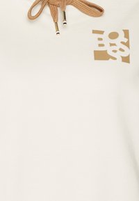 Sudadera color beige claro con un pequeño logotipo marrón en la esquina superior izquierda y cordones ajustables marrones con detalles dorados.