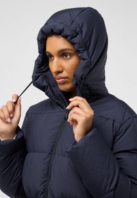 Donkerblauwe pufferjack met een trekkoordkap, rits aan de voorkant en een geruit patroon. De stof lijkt glad en geïsoleerd voor warmte.