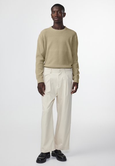 Gebreide trui in beige met ribbelstructuur; gecombineerd met crèmekleurige wijde broek. Donkere schoenen maken de outfit compleet, staande op een neutrale achtergrond.