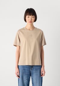 Donna con capelli corti e scuri che indossa una maglietta beige a maniche corte con logo ricamato, abbinata a jeans blu, in piedi davanti a uno sfondo semplice.