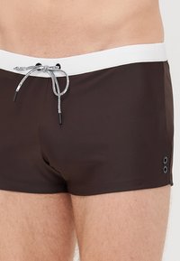 Barna fürdőnadrág fehér húzózsinórral a derékrészen, két fekete füllel az oldalon a biztos illeszkedés érdekében. Sima, rugalmas anyag.