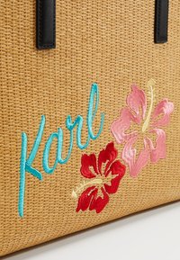 Słomkowa torba na ramię w odcieniu brązowym z czarnymi uchwytami, niebieskim kursywą napisem "Karl" oraz haftowanymi różowymi i czerwonymi kwiatami hibiskusa.