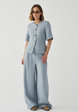 Naine, kes kannab helesinist lühikeste varrukatega nööbitavat pintsakut sobivate laiade pükstega ja pruune mokassiinjalatseid, seisab ühevärvilise tausta ees.