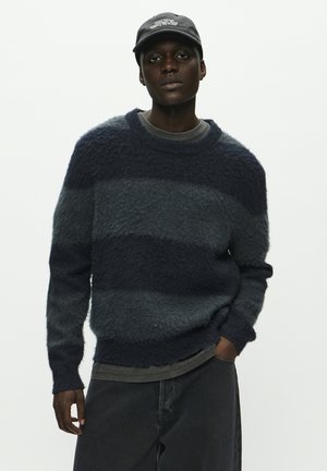 Suéter a rayas en azul marino y gris con una textura suave y cuello redondo. Lleva encima una camisa gris oscura, combinada con jeans oscuros.