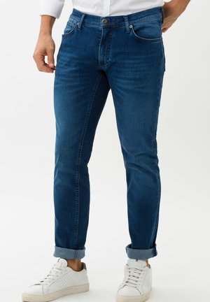 Mężczyzna w ciemnoniebieskich podwiniętych jeansach i białych trampkach, stojący z jedną ręką blisko kieszeni na jednolitym białym tle.