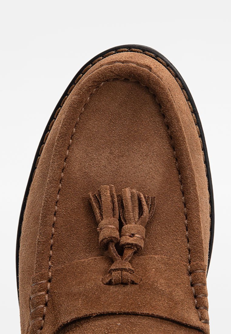 Braune Wildleder-Lederschuhe mit runder Spitze, Quastenakzenten und kontrastierender schwarzer Sohle. Weiche Textur und detaillierte Nähte entlang der Kanten.