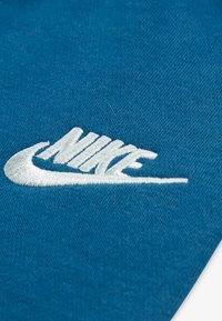 Nikejev logotip iz bele vezenine na teksturirani modri tkanini, s kisikovo oblikovanje, ki poudarja značilno obliko in slog logotipa.