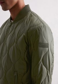 Gequilted bomberjack in matte olijfgroen, met een ribgebreide kraag en manchetten, voorzien van een klein merklabel op de mouw en een zippocket aan de zijkant.