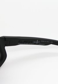 Čierne slnečné okuliare s matným povrchom, uhlovým dizajnom rámu a sivými sklami. Obsahujú text na obrubách, ktorý uvádza značku a UV ochranu.