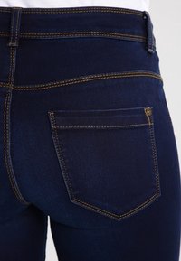Mörkblå denimjeans med en slät textur, prydda med gyllene sömmar och en bakficka på höger sida. Enkelt design, avslappnad passform.