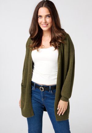 Cardigan vert olive avec une coupe décontractée, des poignets côtelés et une ouverture à l'avant. Porté sur un débardeur blanc et associé à un jean bleu et une ceinture noire.