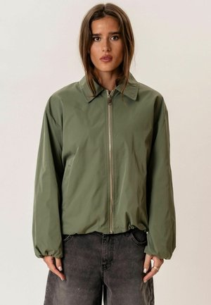 Fiatal nő olívazöld, cipzáras, galléros és bő ujjú dzsekit visel, sötétszürke farmerrel, egyszínű háttér előtt állva.