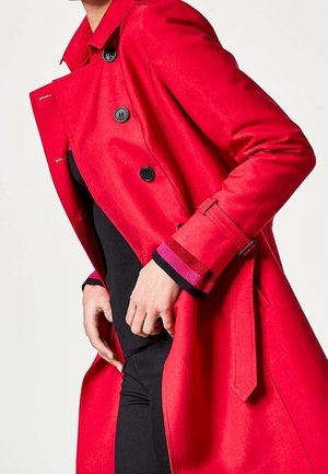 Kvinde iført en knaldrød, dobbeltradet trenchcoat med sorte knapper over et sort outfit, hænder justerer frakkens revers.