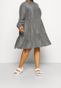 Grijze, jurk met lange mouwen en een knoopsluiting kraag en een tiered rok. Gepaard met witte sandaaltjes met bandjes. Zachte textuur en een ontspannen pasvorm.