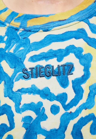 Κοντινό πλάνο ενός πουκαμίσου με μπλε και κίτρινα αφηρημένα σχέδια και τη λέξη "STIEGLITZ" κεντημένη με μαύρο νήμα στο στήθος.