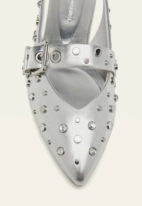 Scarpa con tacco alto argento a punta, decorata con borchie circolari e strass, dotata di una larga cinghia per la caviglia con fibbia.